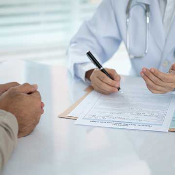 A person in a white coat does paperwork and talks to another person