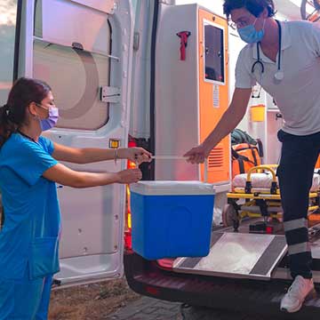 A person in an ambulance hands a cooler to an individual wearing blue scrubs